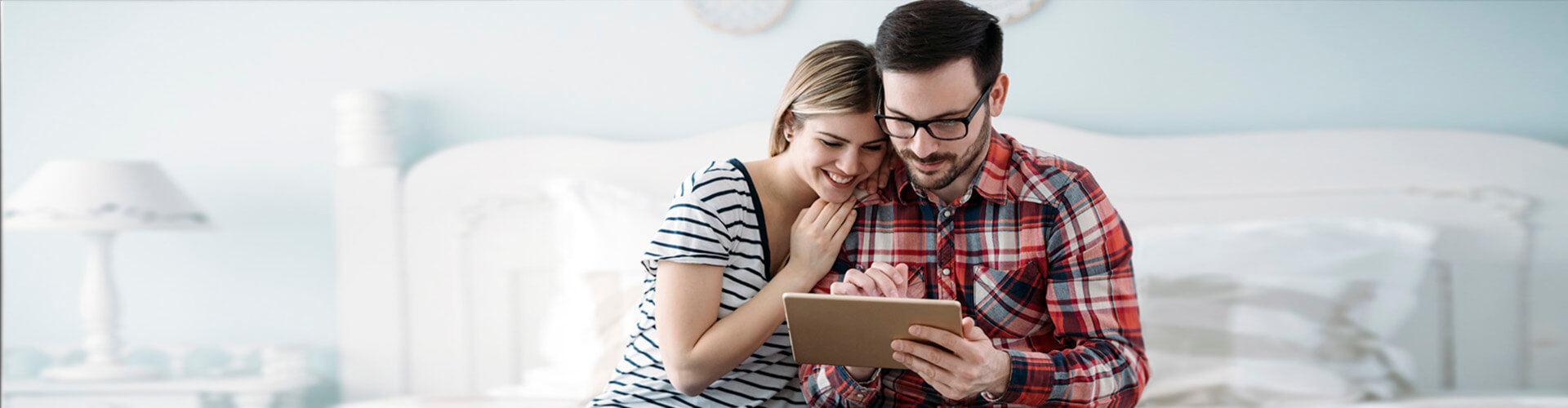 Programa te lo mereces nomina y pension - Pareja sentada en su cama en la habitacion con la tablet en la mano sonriendo Programa te lo mereces nomina y pension - Pareja sentada en su cama en la habitacion con la tablet en la mano sonriendo