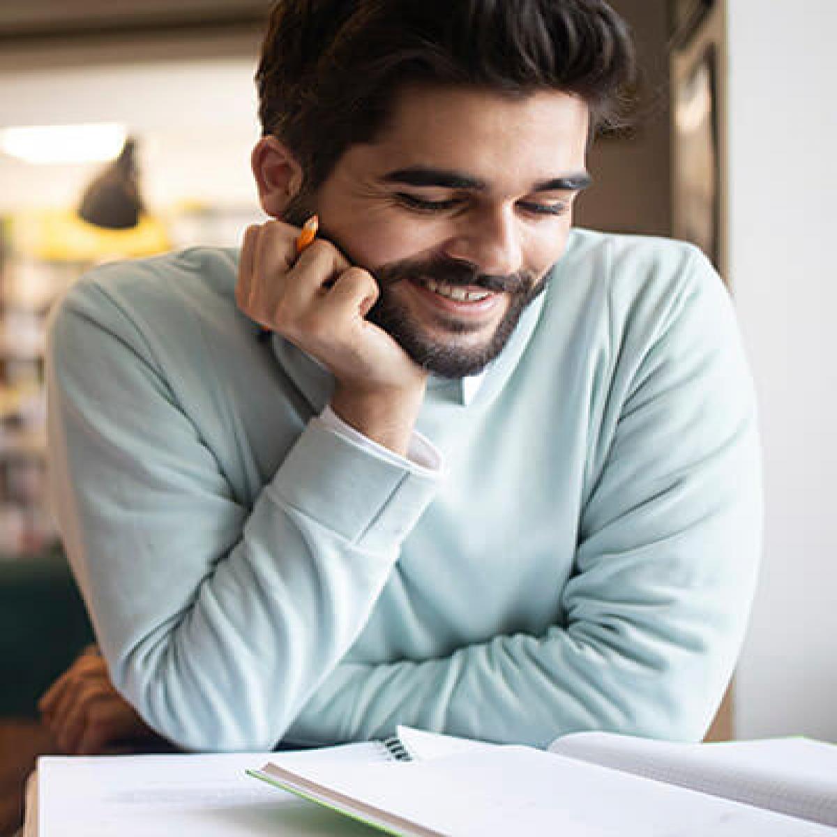 ¿Dónde quieres buscar empleo?  - Estudiante feliz y sonriendo preparando un examen en la biblioteca rodeado de libros