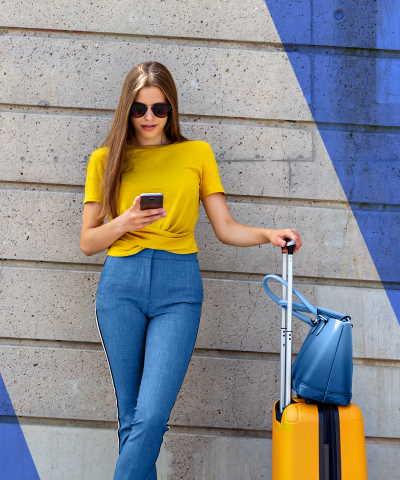 Momento Viajar al extranjero - mujer-joven-con-camiseta-amarilla-y-maleta-usando-su-movil-apoyada-contra-una-pared-azul-y-gris - Caja Rural de Navarra
