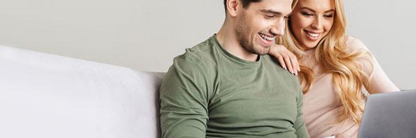 Recomendador de Tarjetas - Pareja joven y feliz sonriendo mientras navegan con el portatil sentados en el sofa de su casa con una tarjeta en la mano Recomendador de Tarjetas - Pareja joven y feliz sonriendo mientras navegan con el portatil sentados en el sofa de su casa con una tarjeta en la mano